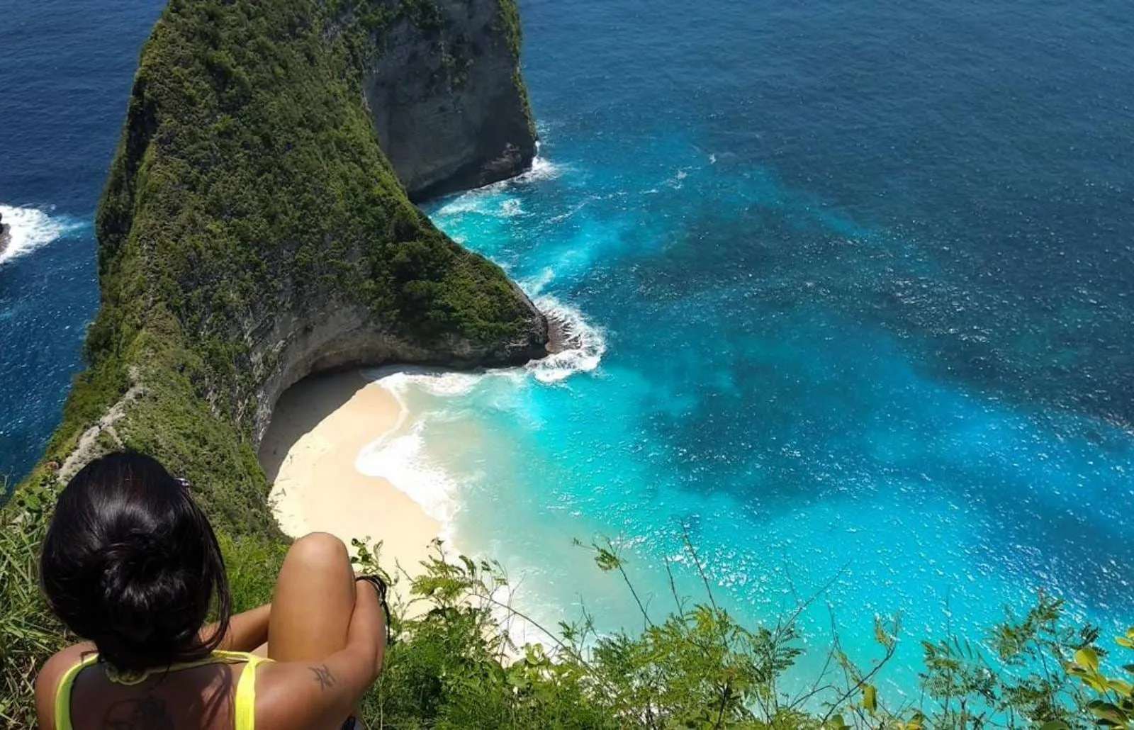 Frau sitzt am Klippenrand und blickt auf den türkisfarbenen Strand und die steile Bucht von Nusa Penida.